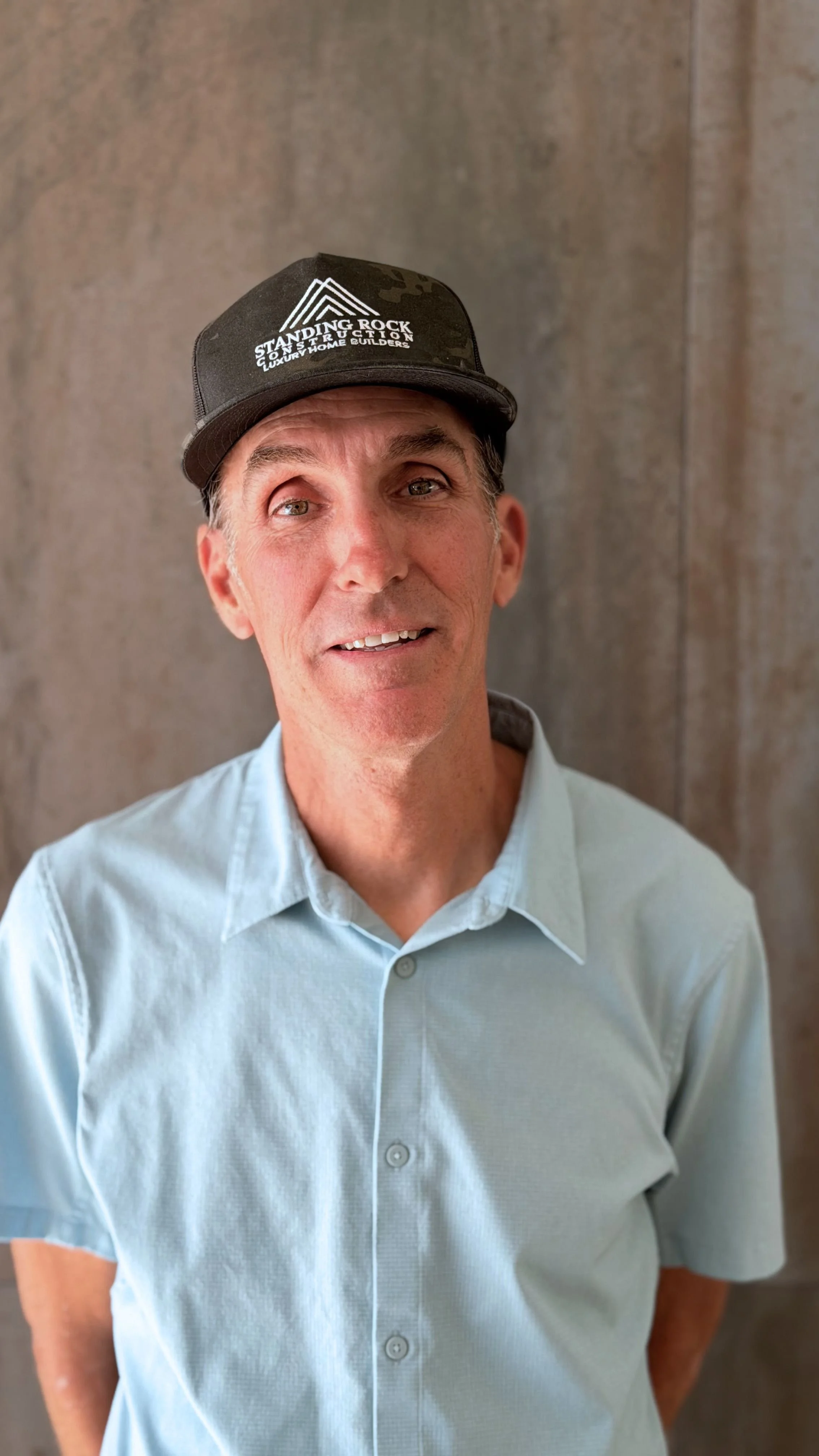 A man with light skin and gray hair, wearing a light blue dress shirt and a black trucker hat with white logo and text, standing in front of a plain, textured background.