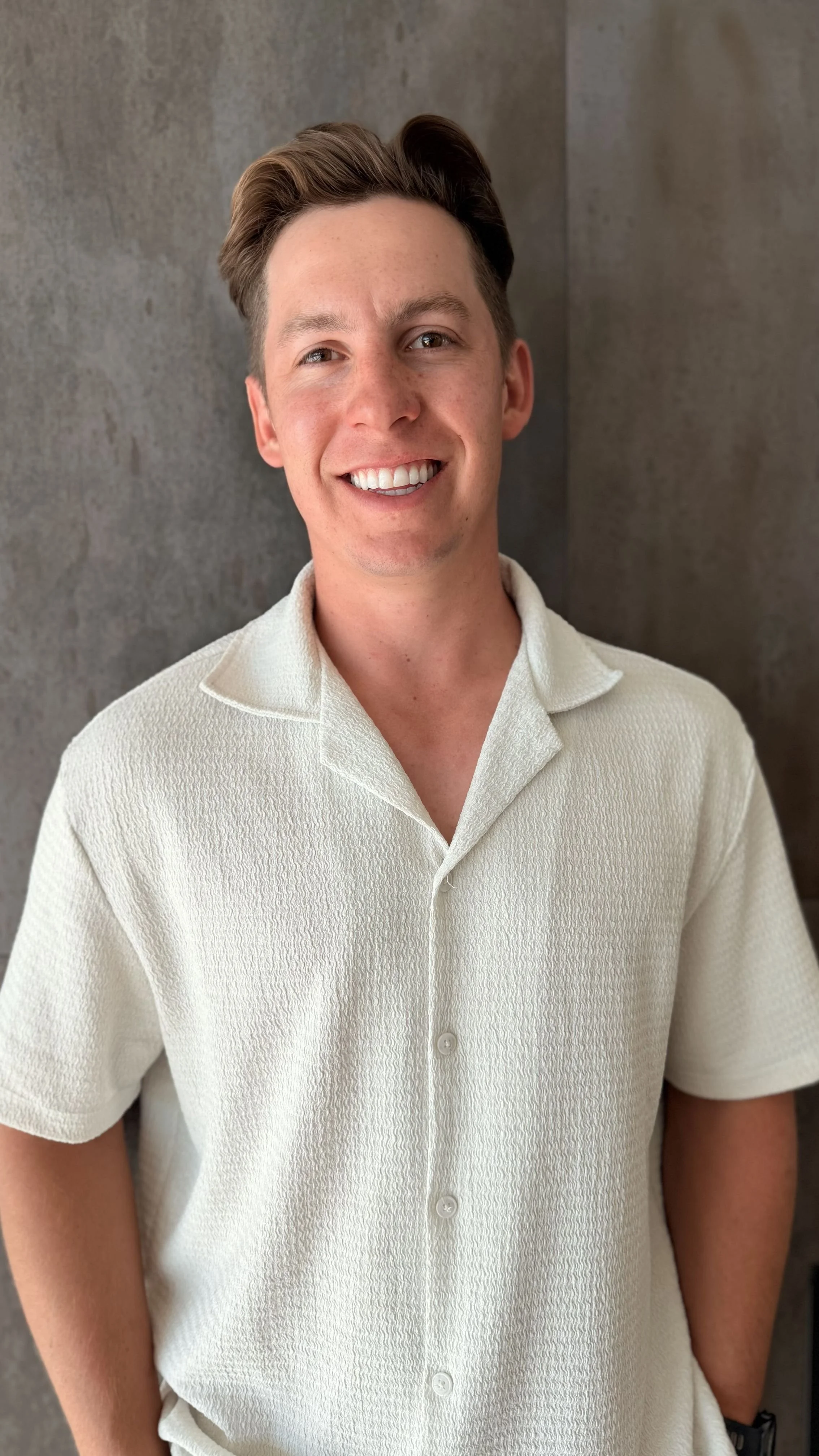 Young man with light skin and brown hair, smiling, wearing a cream-colored short-sleeve button-up shirt, standing against a gray wall.