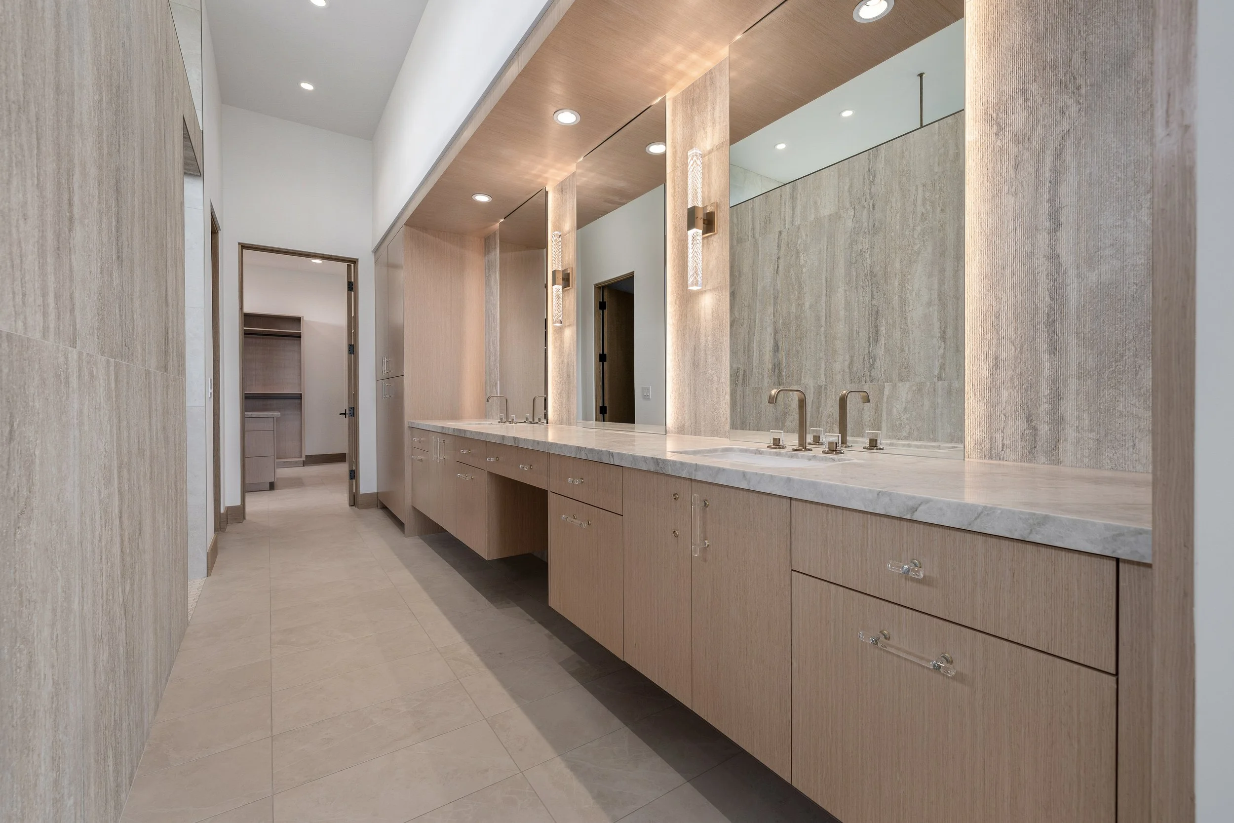 Modern bathroom with beige stone walls and flooring, wooden cabinets with marble countertops, and sleek fixtures under recessed lighting.
