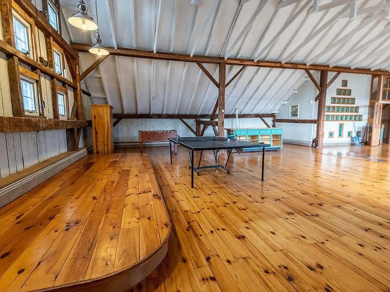 Spacious attic room with wooden floors, sloped ceiling, and exposed beams, furnished with a ping pong table and small storage units.