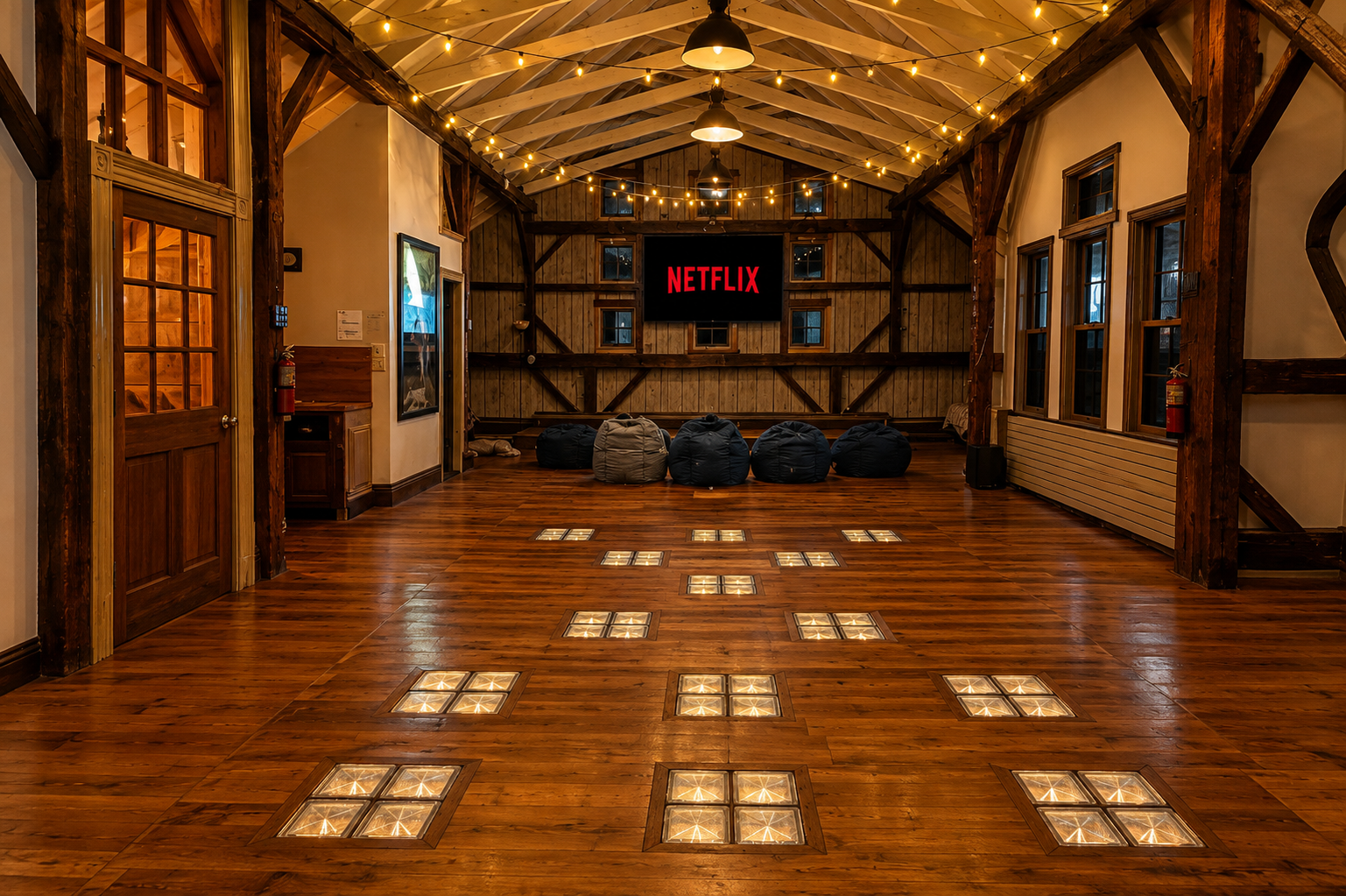 Interior of a rustic event space with wooden floors, string lights on the ceiling, and a large TV screen displaying the Netflix logo, surrounded by bean bags for seating.