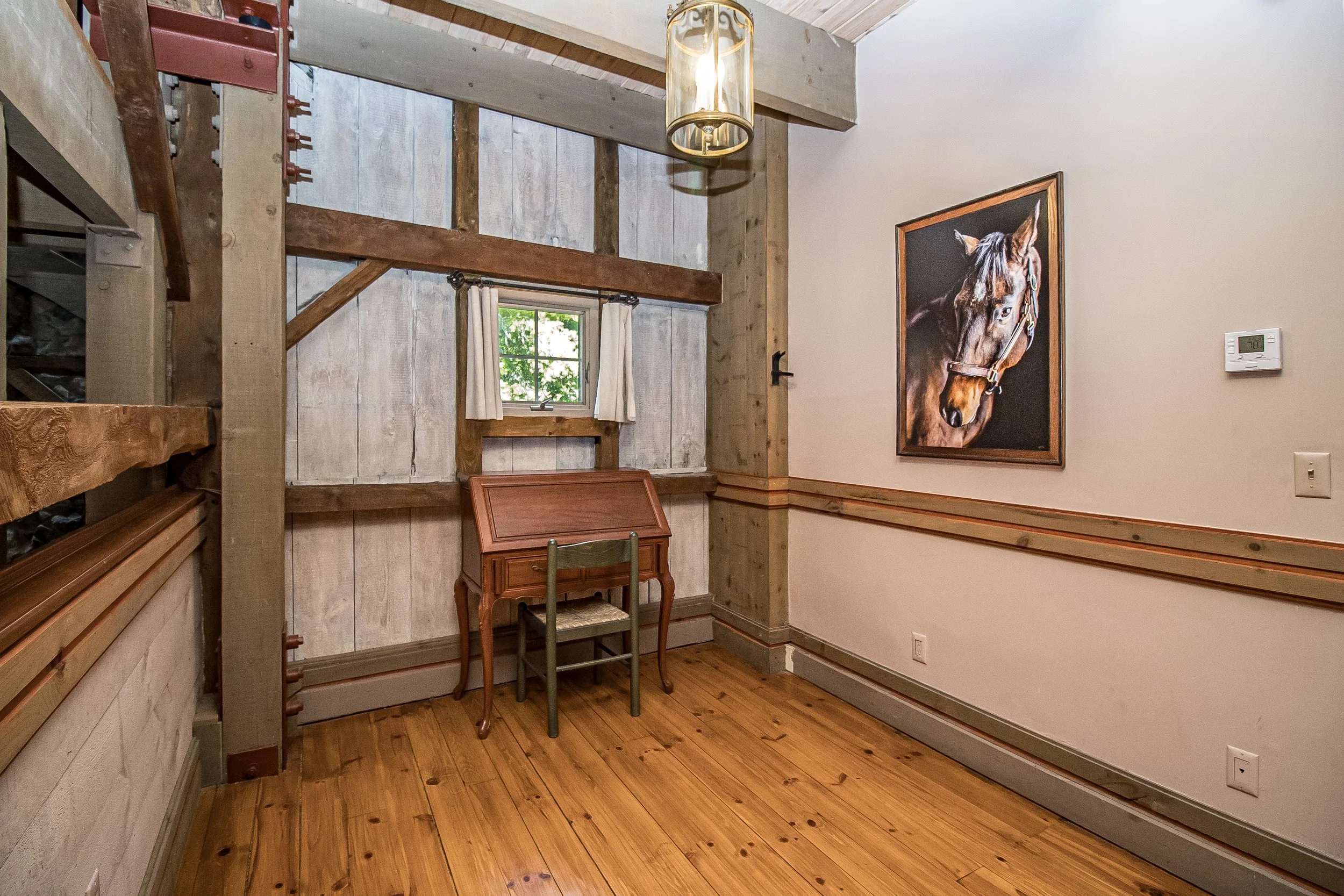 A corner of a rustic room with wood-paneled walls, a window with white curtains, a wooden desk with a chair, a framed photograph of a horse, a vintage light fixture, and a thermostat on the wall.