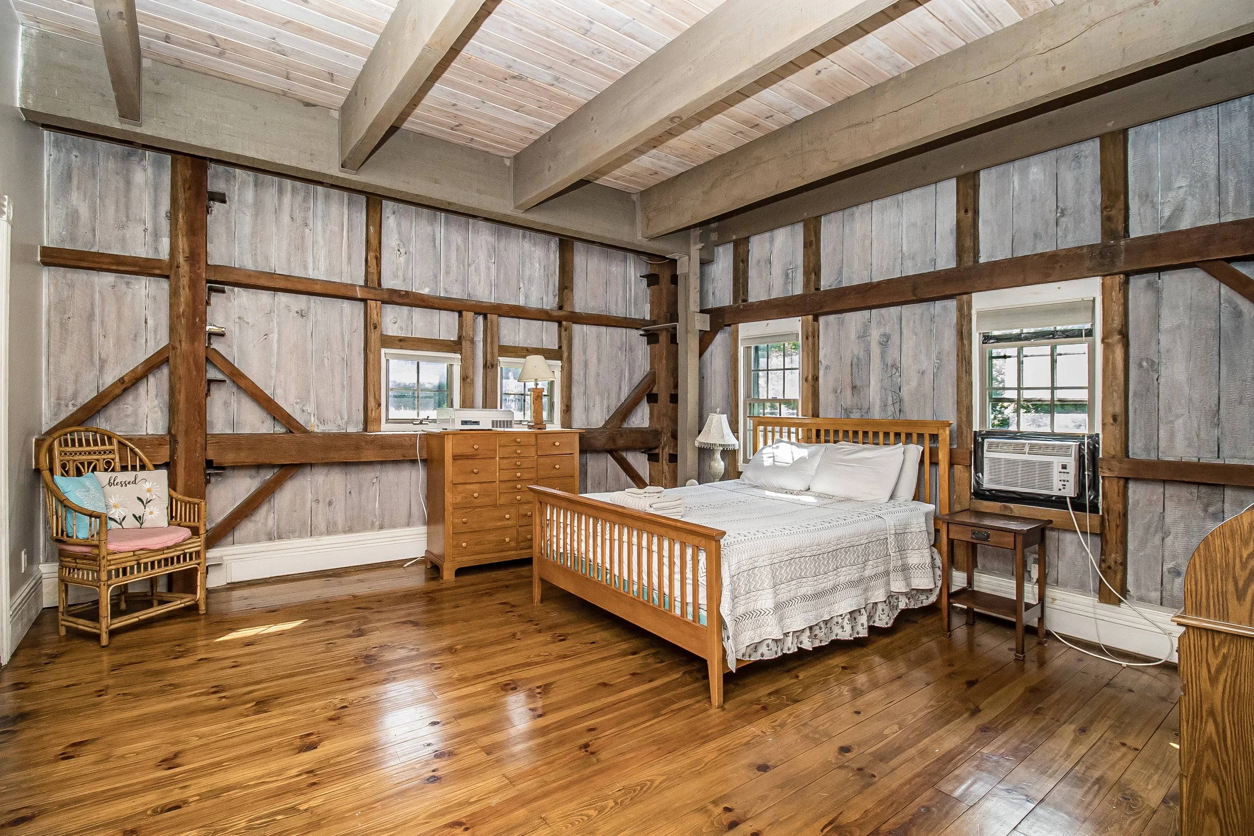 Interior of a bedroom with wooden walls and ceiling beams, a wooden bed with white bedding, a wooden dresser, a wicker chair with cushions, windows with blinds, and wood flooring.