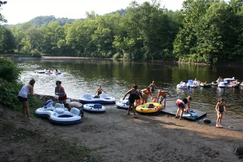 People preparing to float on inner tubes and rafts at a river beach, with trees and hills in the background.
