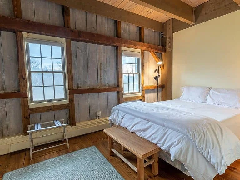 A cozy bedroom with wooden walls, two windows, a bed with white bedding, a small wooden bench at the foot of the bed, and a wall-mounted reading lamp.