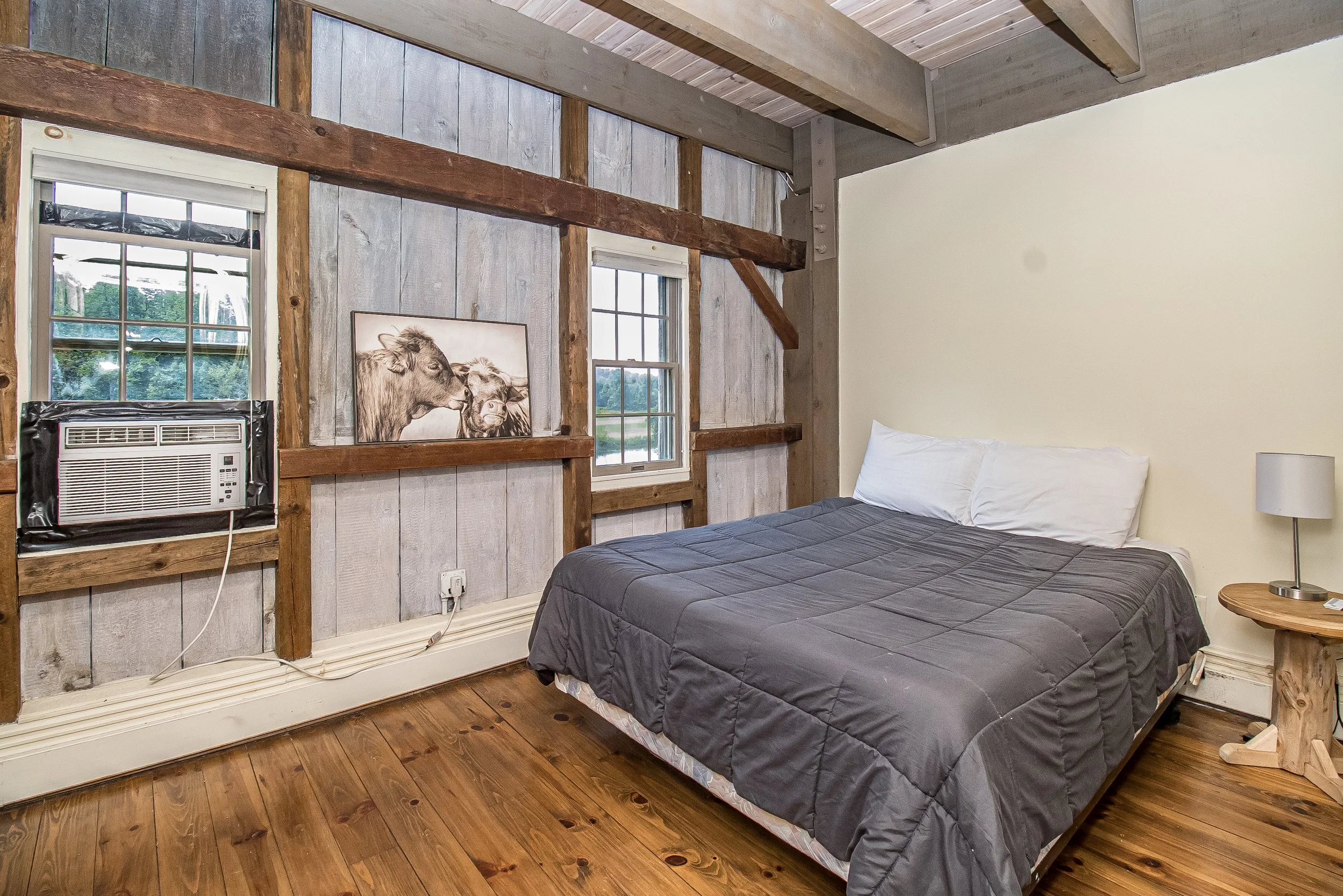 A rustic bedroom with wooden walls and ceiling, featuring a bed with a dark gray quilt and white pillows, a round wooden side table with a lamp, and two windows. There is a framed artwork of two cows on the wall, and a window air conditioning unit below one of the windows.