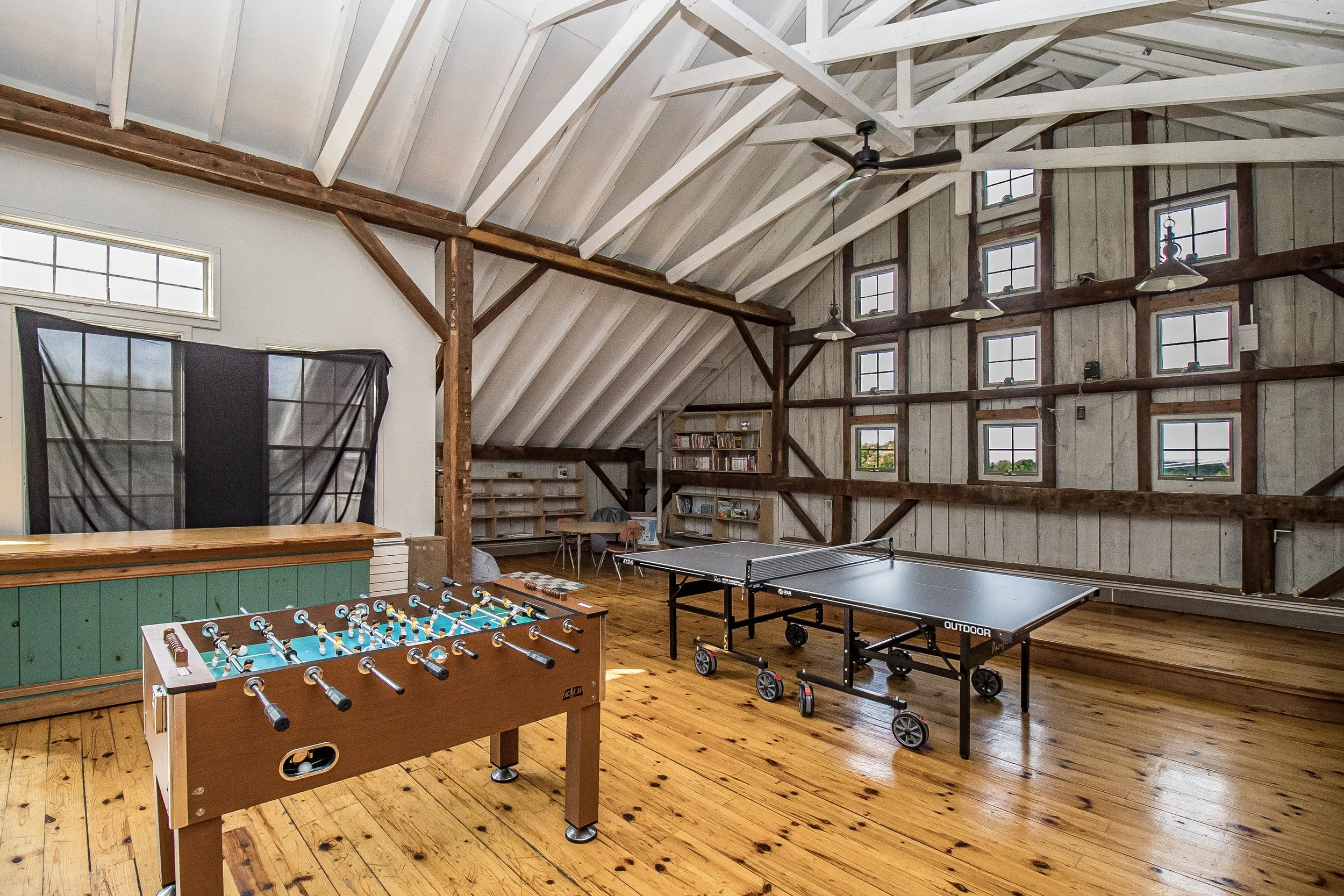 The image shows a spacious indoor game room with wooden floors and a high ceiling with white beams and a ceiling fan. There is a foosball table in the foreground, a ping pong table towards the back, and a foosball table on the right. The walls are partly wooden and have multiple small windows that let in natural light.