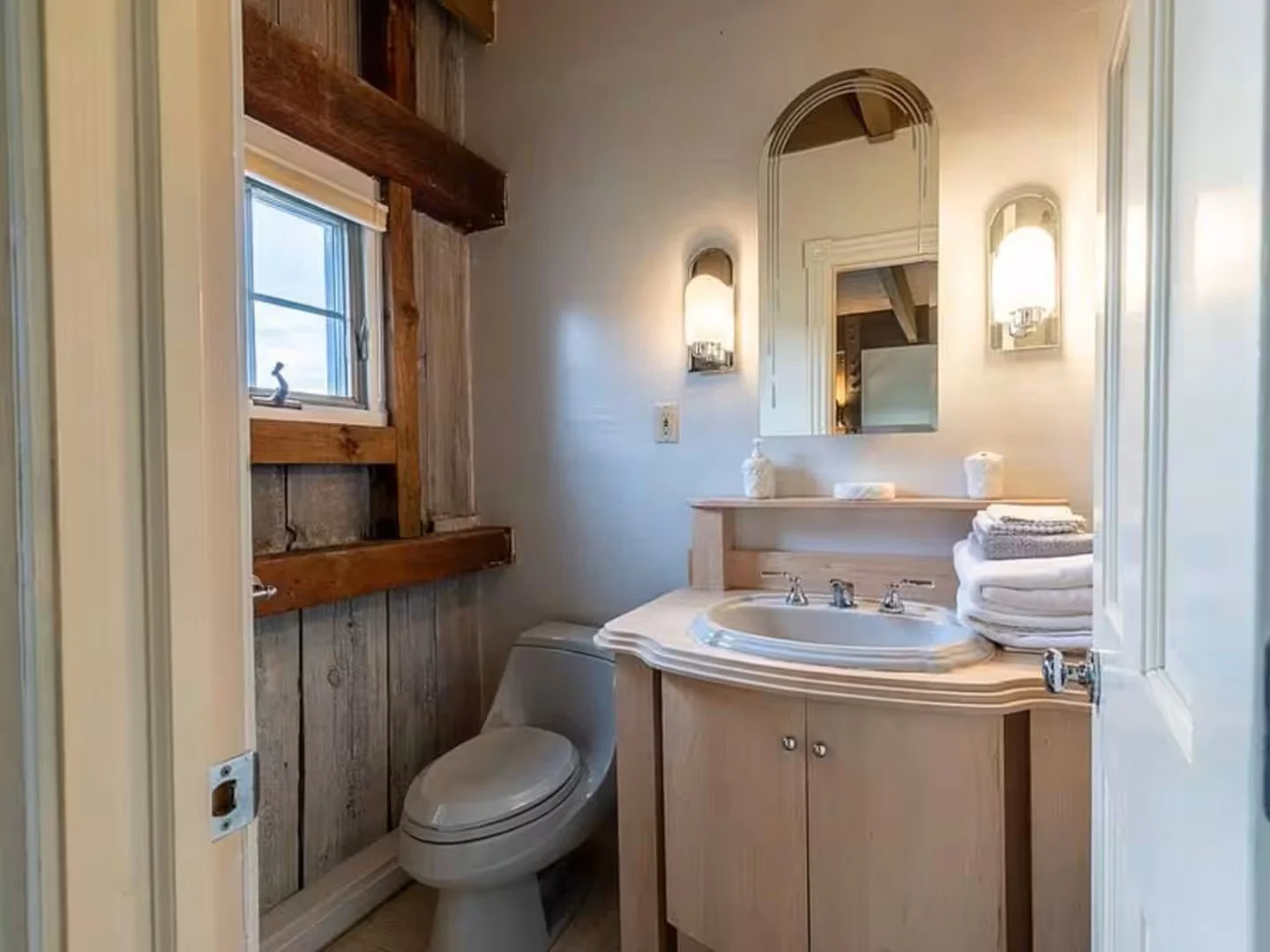 A rustic bathroom featuring a beige vanity with a curved countertop and a built-in sink, topped with neatly folded towels and a soap dispenser. Above the vanity is a mirror flanked by two wall-mounted light fixtures. To the left, there is a small window with a Roman shade and wooden beams on the wall. The toilet is next to the vanity, and the walls have a mixture of textured wood and painted surfaces.