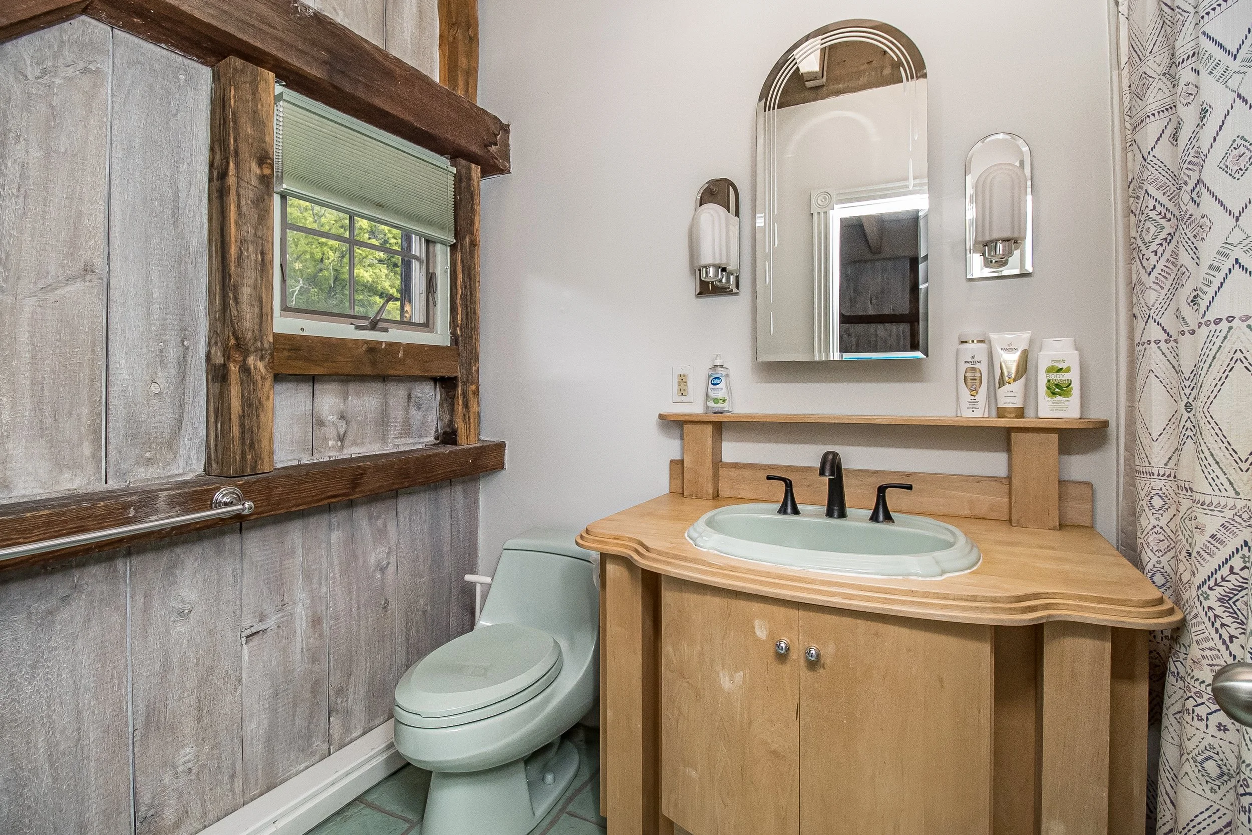 A rustic bathroom with a small window with blinds, a mirror above a wooden counter, a green toilet, and various toiletries on the counter.