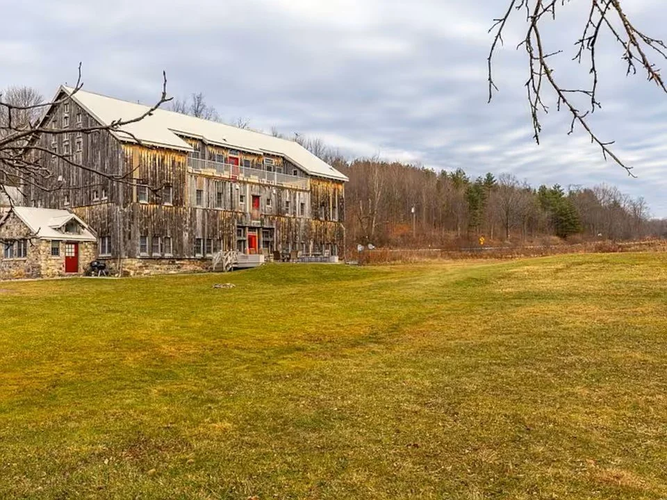 A rustic multi-story barn with a weathered wood exterior, red doors, and a stone foundation, situated on a large grassy field with leafless trees and a cloudy sky in the background.