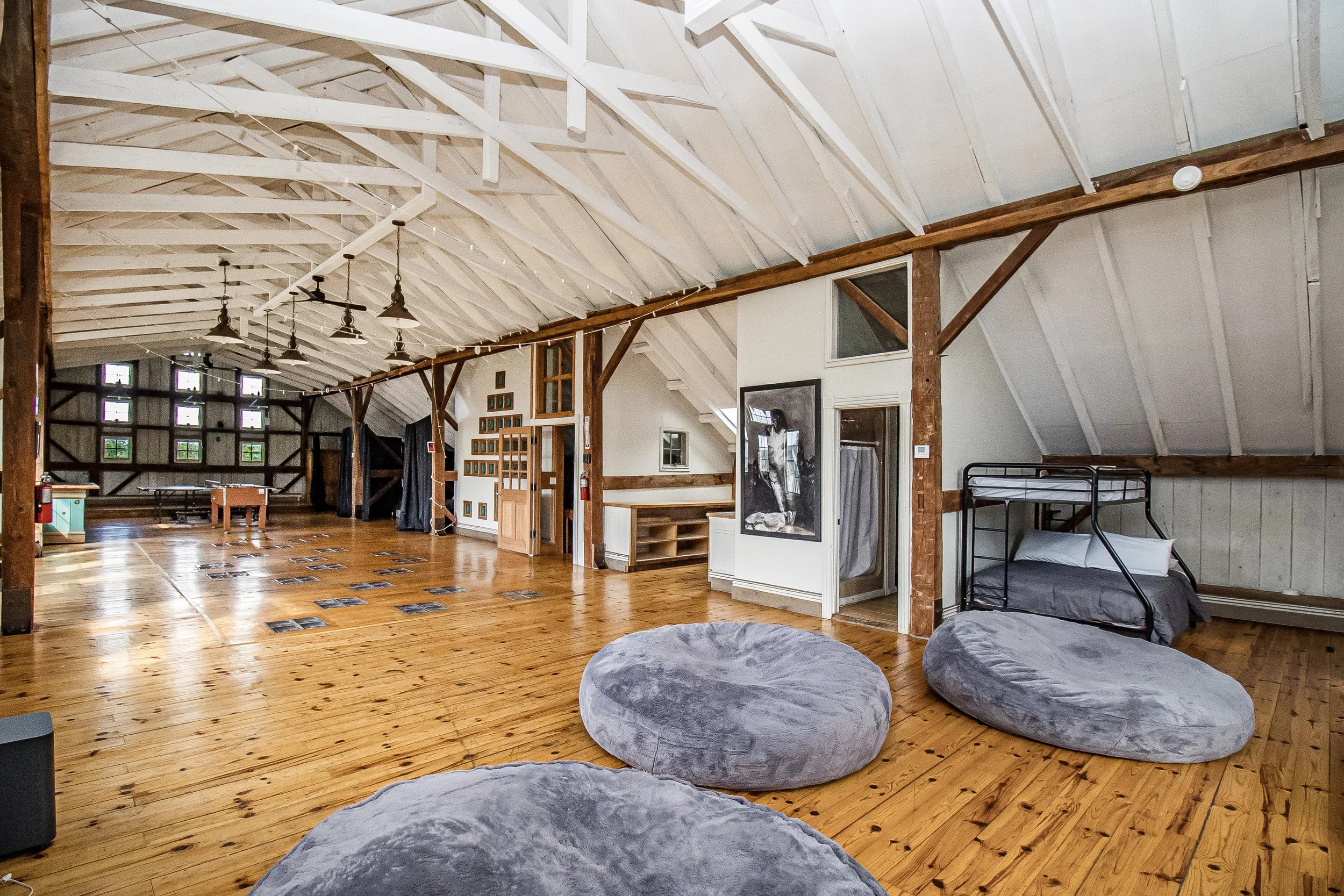 A spacious room with exposed wooden beams and a sloped ceiling, wooden flooring, multiple windows, hanging pendant lights, a bunk bed, artwork on the walls, and large gray bean bag chairs.