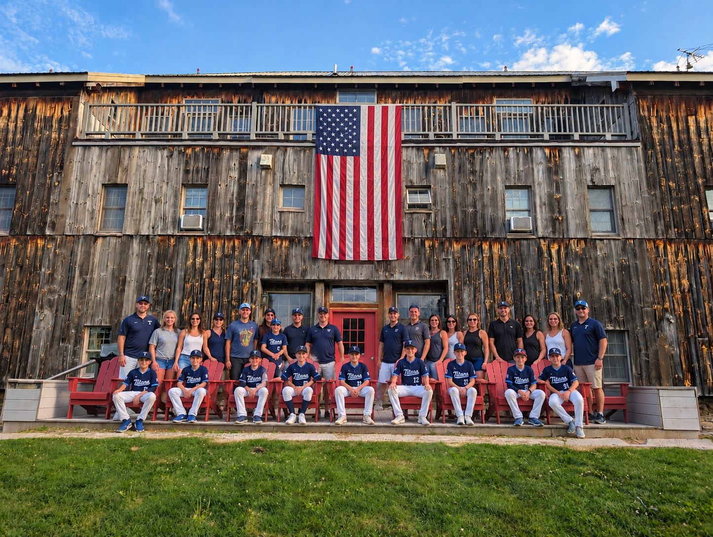 A group of Cooperstown dreams park baseball players in blue uniforms sitting on red benches in front of a rustic wooden building with an American flag hanging on it, under a partly cloudy sky.