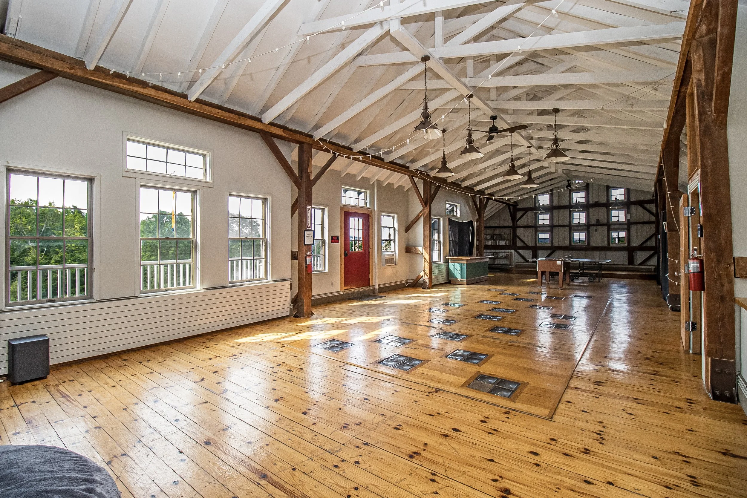 Spacious room with high vaulted ceiling, large windows letting in natural light, wooden floor, hanging ceiling fans, and string lights, featuring a rustic and modern decor.