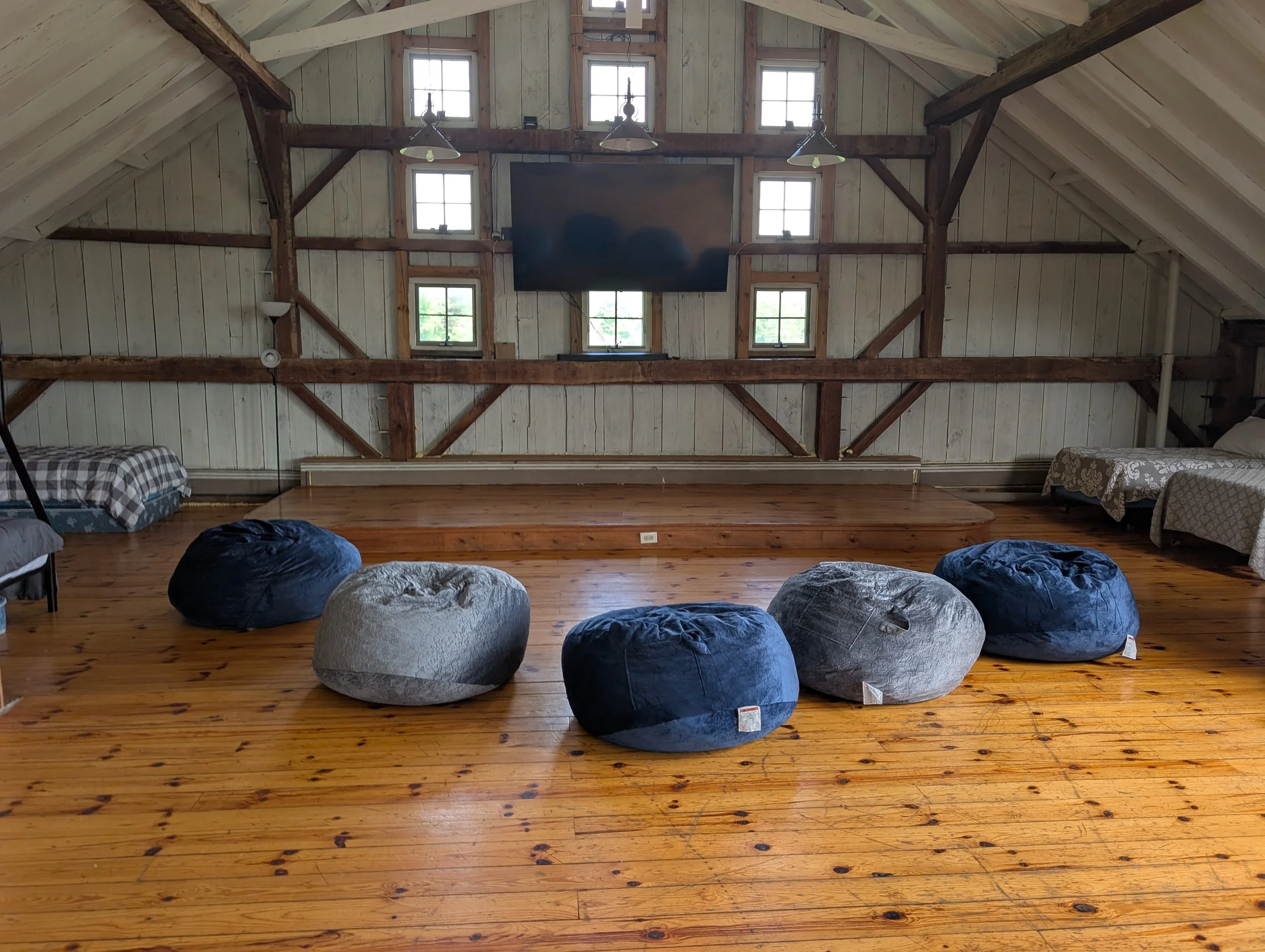 Wooden-floored room with six bean bag chairs, a wall-mounted flat screen TV, and multiple small windows in a barn-like structure.