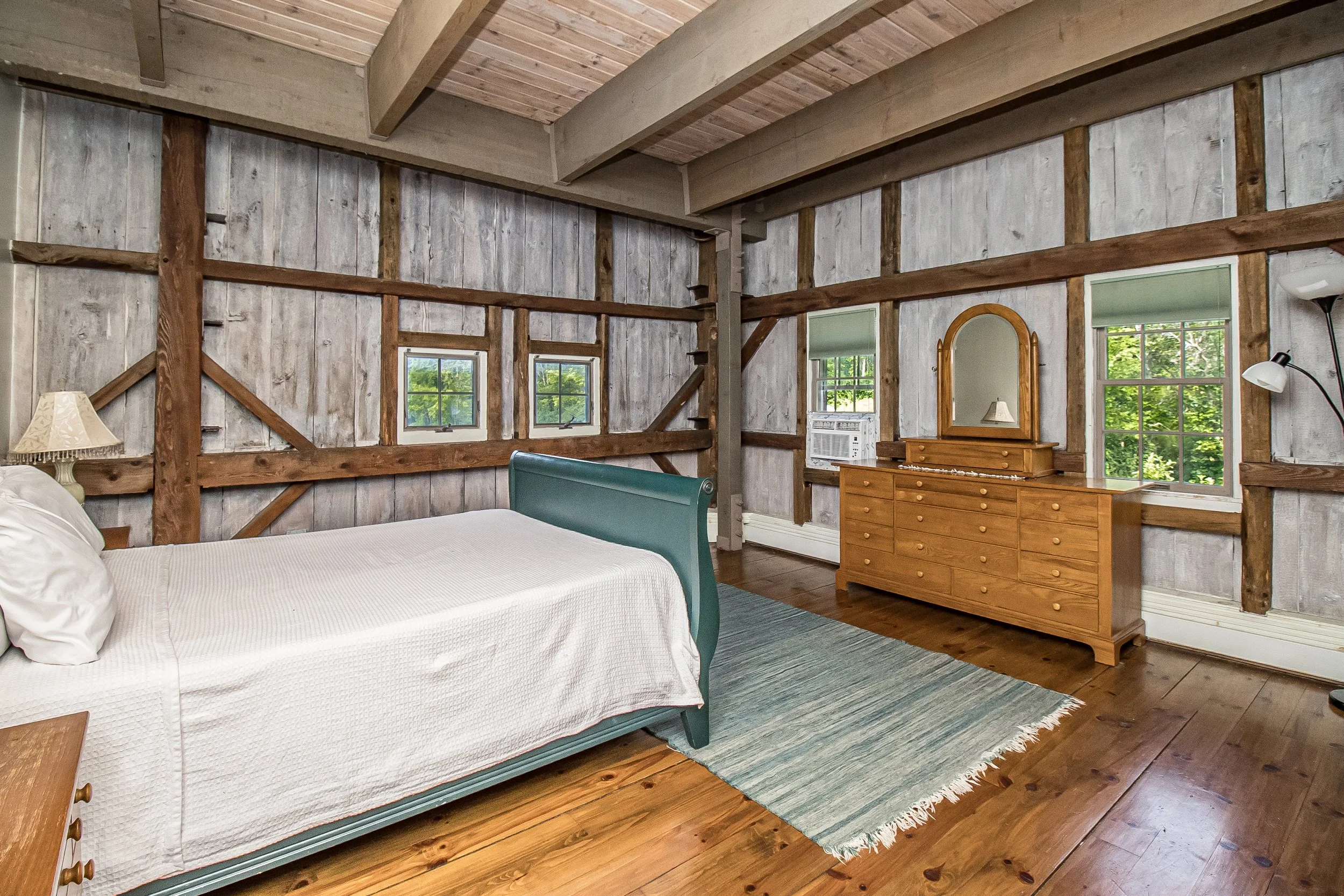A rustic bedroom with wooden walls and ceiling, featuring a bed with white bedding, a wooden dresser with a mirror, and windows showing greenery outside.