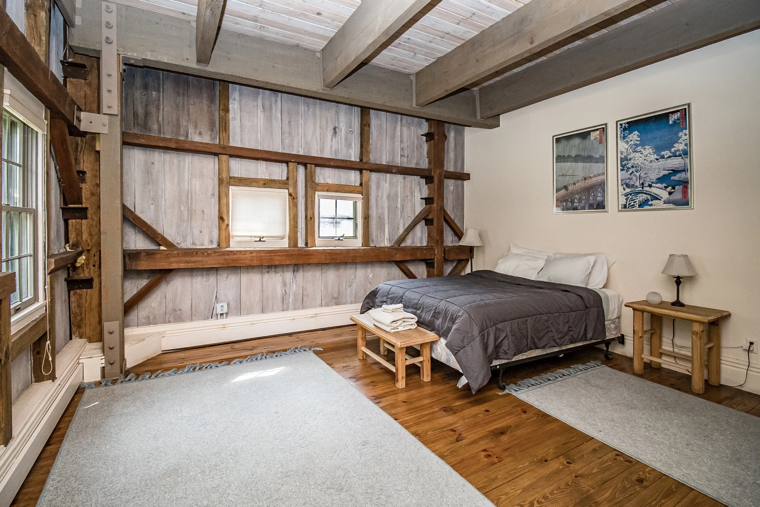 A bedroom with a rustic design featuring wooden beams, a wood-paneled wall, two small windows, a made bed with a gray comforter, a wooden bench at the foot of the bed with folded towels, two small wooden nightstands with lamps, a framed artwork on the wall, and a mix of hardwood flooring and a rug.