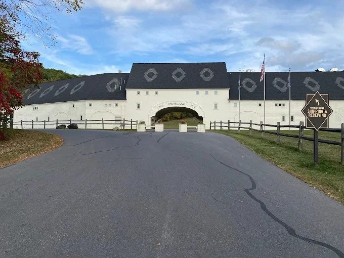 Large white building with a black roof and an arched entrance, surrounded by a wooden fence, with a road leading up to it and a sign that says 'Shipping & Receiving'.