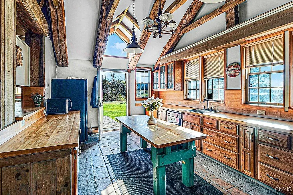 Sunlit rustic kitchen with wooden cabinets, a green kitchen island, a row of windows with blinds, and exposed wooden beams on the ceiling.