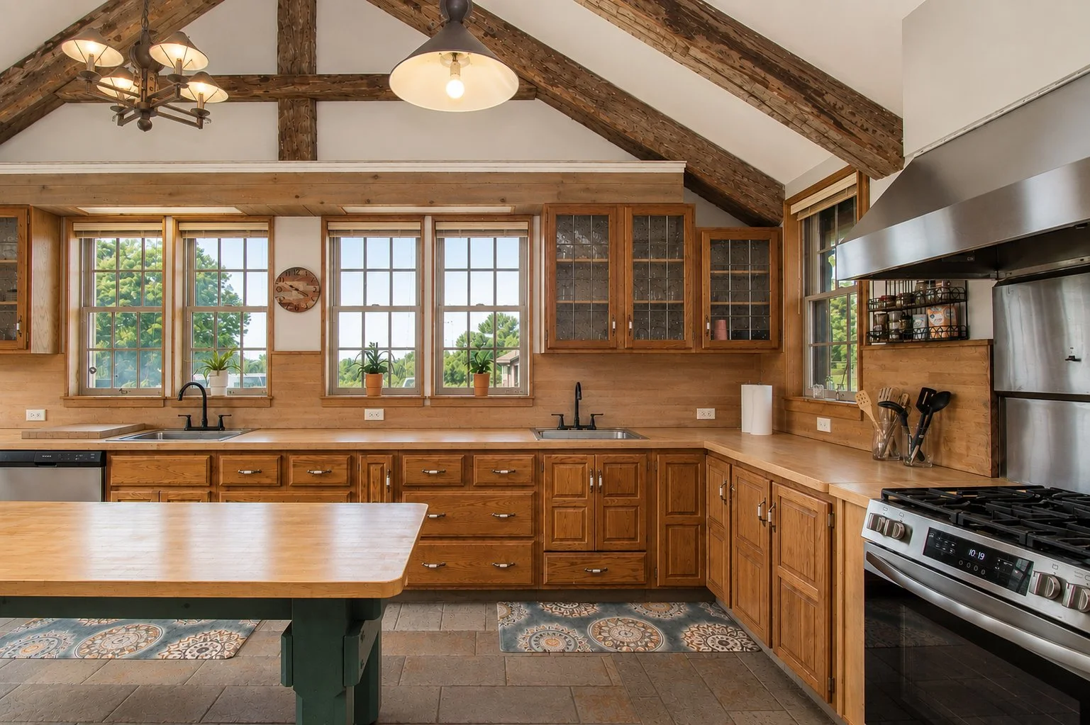 A spacious kitchen with wooden cabinets, a center island, three large windows with plants, and a sloped wooden ceiling with exposed beams.