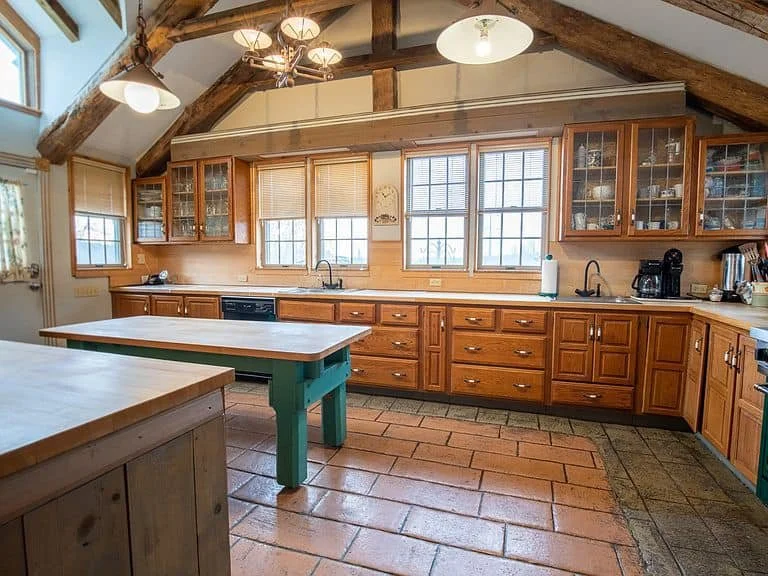 Kitchen with wooden cabinets, tile flooring, multiple windows, and rustic wooden beams on ceiling. Includes kitchen appliances and a green kitchen island.