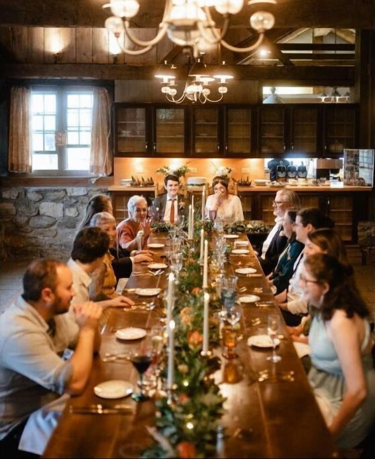 People gathered around a long dining table with candles and a floral centerpiece, enjoying a meal in a rustic, warmly-lit room with wooden walls and ceilings.