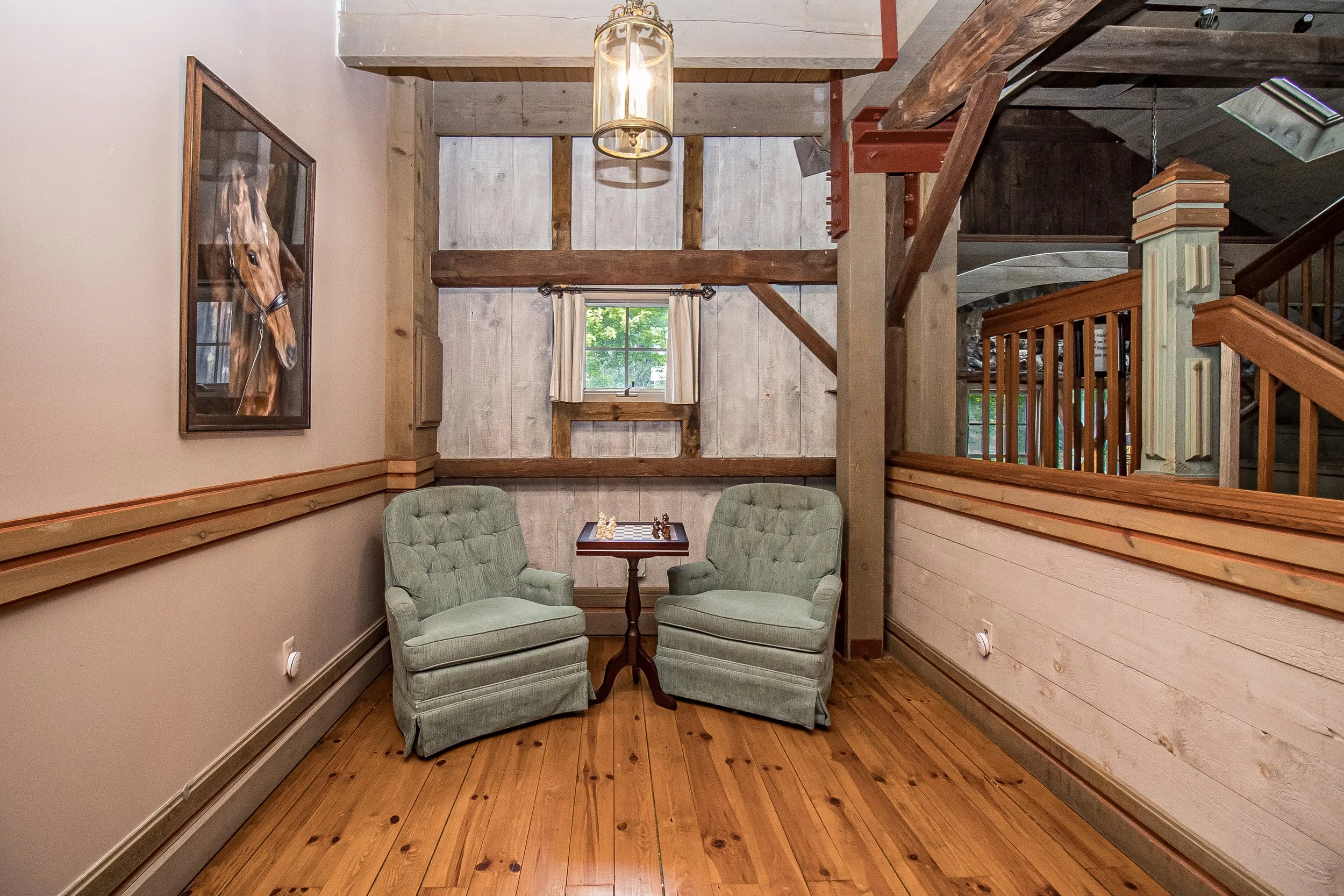 A cozy corner with two green armchairs facing each other, a small wooden table with a chessboard between them, in a room with wooden floors, paneled wooden walls, and a window with white curtains. A large framed horse portrait hangs on a beige wall to the left.