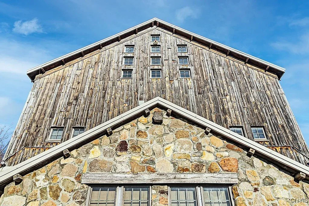 Close-up view of a rustic, weathered barn with a stone base and wooden upper structure, set against a bright blue sky.