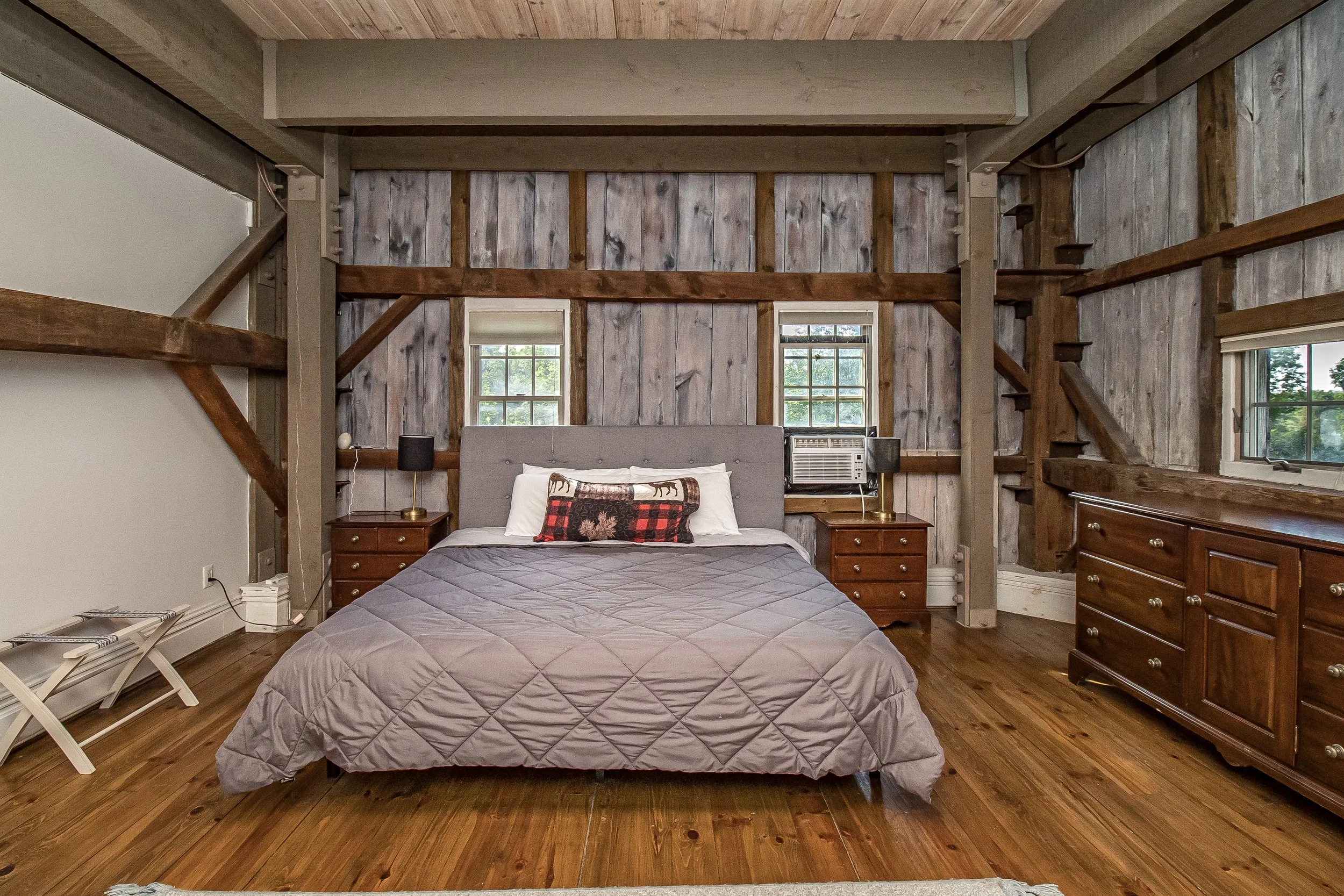 The image shows a bedroom with wood-paneled walls and hardwood floors. There is a bed with a gray quilted bedspread and decorative pillows, flanked by two wooden nightstands with black lamps. Behind the bed, the wall features exposed wooden beams and two windows with a view of trees outside. To the right, there is a large wooden dresser, and on the left side, a white folding chair and some stacked magazines are visible.