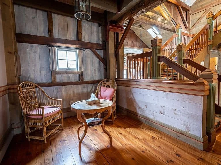 A cozy corner with two wicker chairs with pink cushions, a small wooden table with a tissue box, and a tiny window on a rustic wooden wall inside a cabin or wood-paneled house.