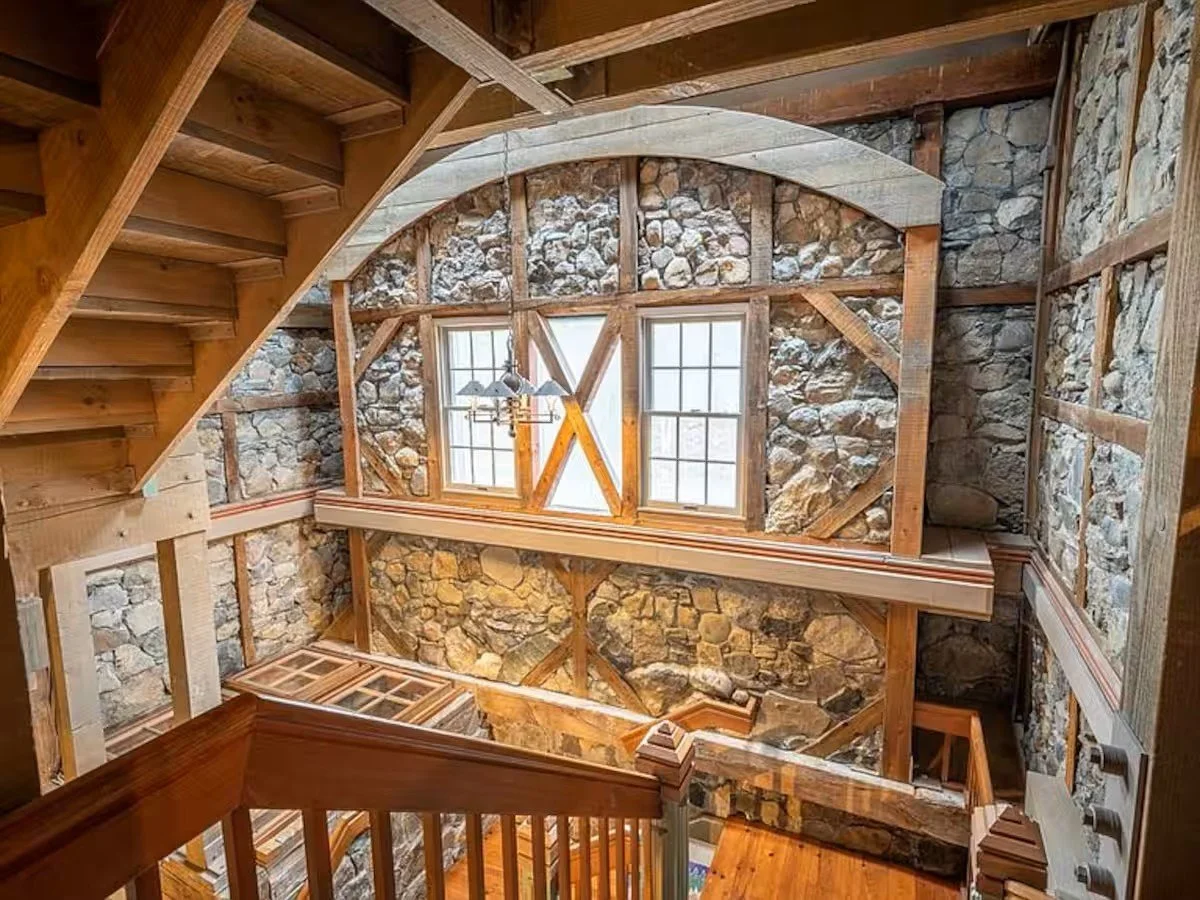 Interior view of a multi-story house with exposed stone walls, wooden beams, and staircases, showcasing a rustic architectural style.