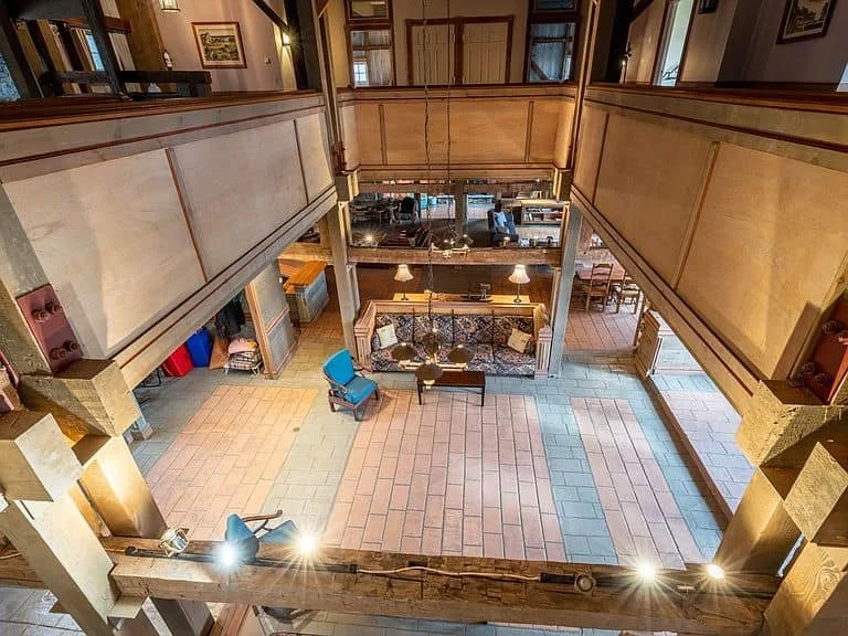 A spacious, multi-level interior with a central open area. The ground floor features tile flooring with different colors, a blue chair, and a vintage-style sofa in the background. The upper level has wooden railings and is visible from above. The roo
