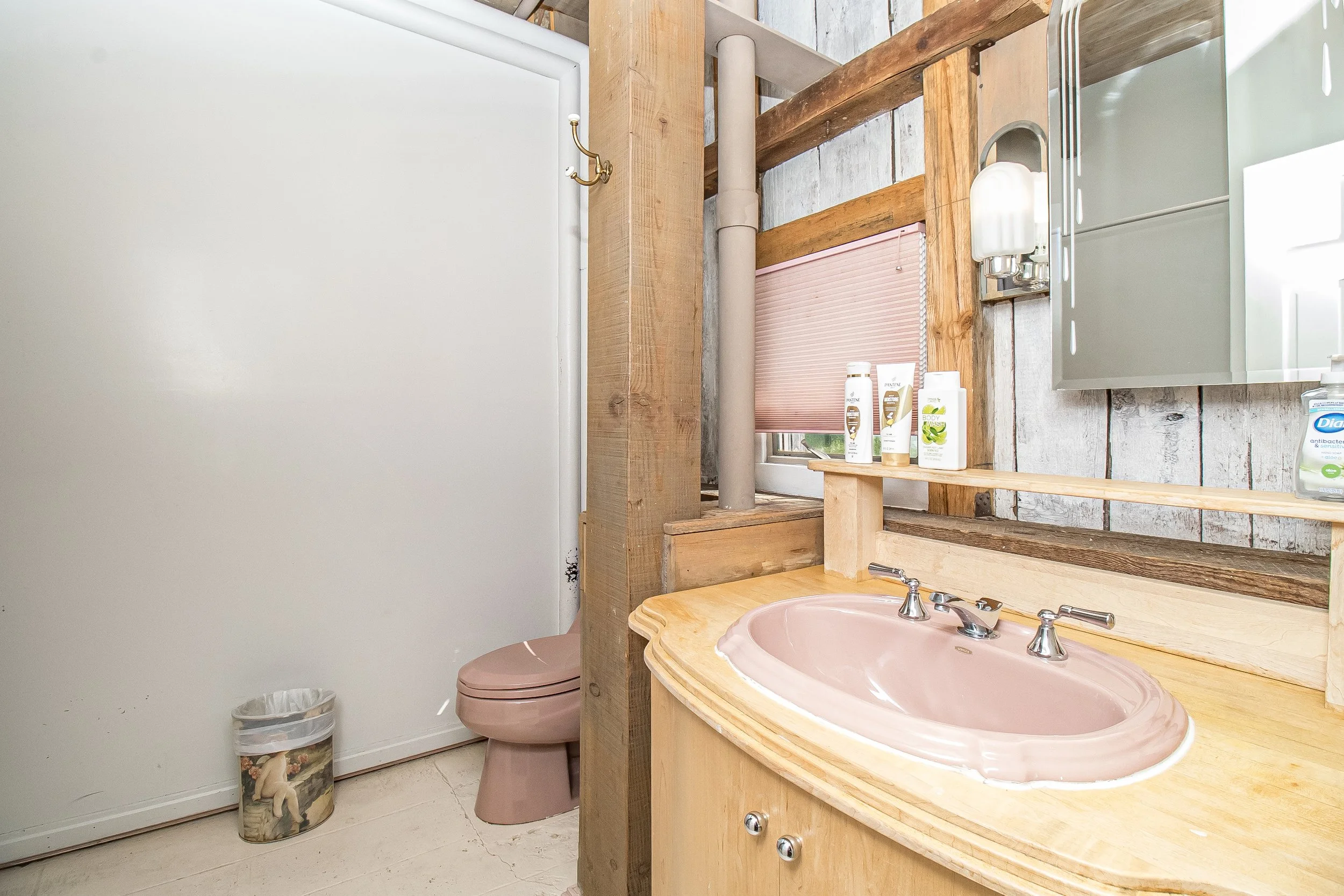 A rustic bathroom with a pink sink, a wall mirror, and a wooden countertop. There are bottles of soap or lotion on the counter, a pink toilet, and a trash bin. The wall has a mix of wood and concrete textures, a small window with a blinds, and a drainage pipe.