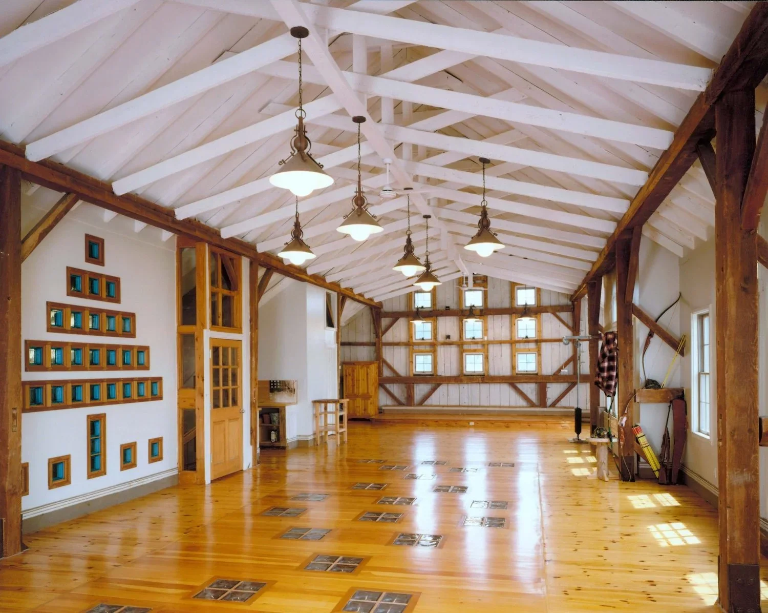The rustic hayloft with wooden floors, white vaulted ceiling with exposed beams, hanging light fixtures, and multiple small windows on the back wall allowing natural light.