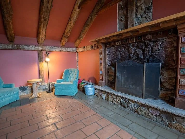 Living room with pink walls, exposed wooden beams, a large stone fireplace with a wooden mantel, blue upholstered chairs, a small round side table, a standing lamp, and baskets near the fireplace.