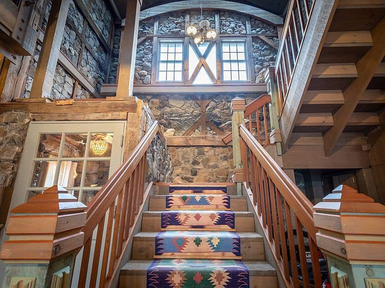 Interior staircase with colorful patterned runner rugs, wooden railings, stone wall, large windows, and a chandelier at the top.