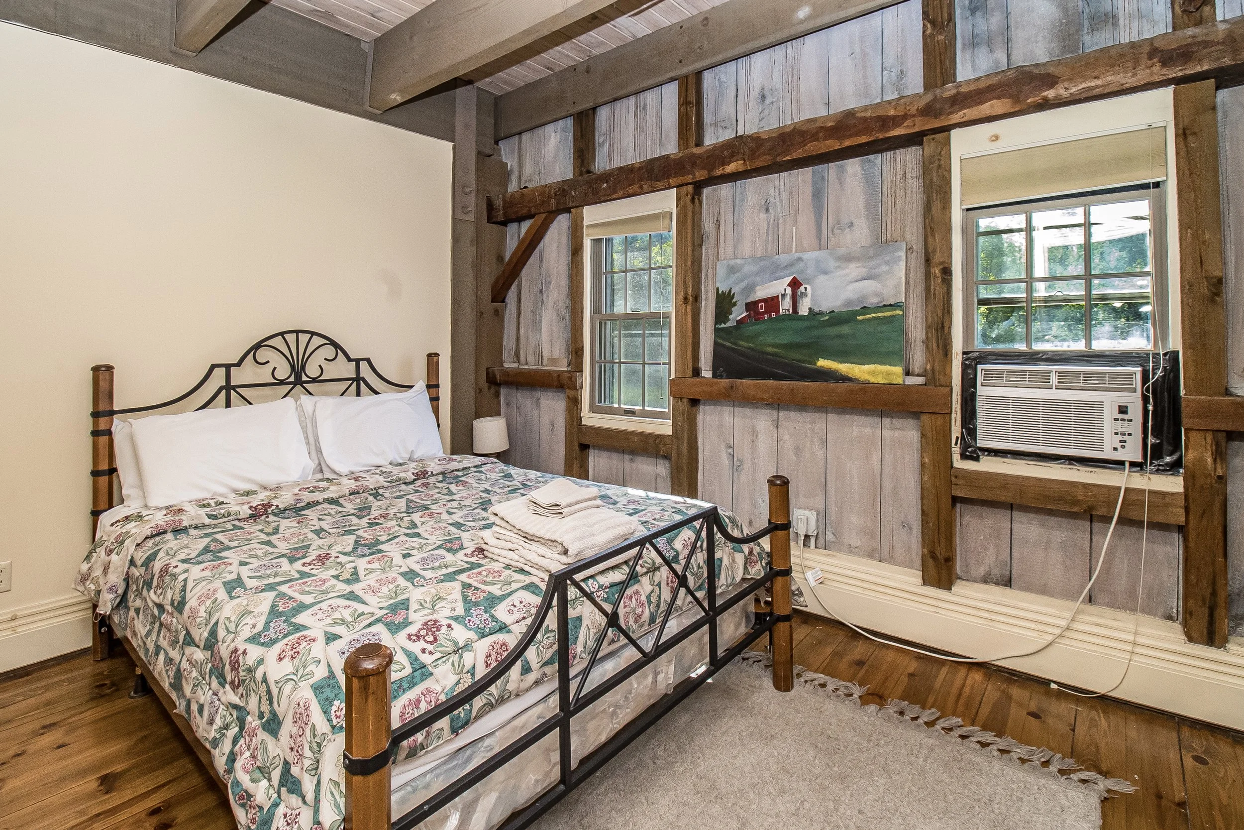 A cozy bedroom with a metal bed frame, a floral bedspread, and two pillows. There is a small lamp on a white bedside table. The room has rustic wood-paneled and partially painted walls, with two windows, one showing a barn painting, and an air conditioning unit under the right window.
