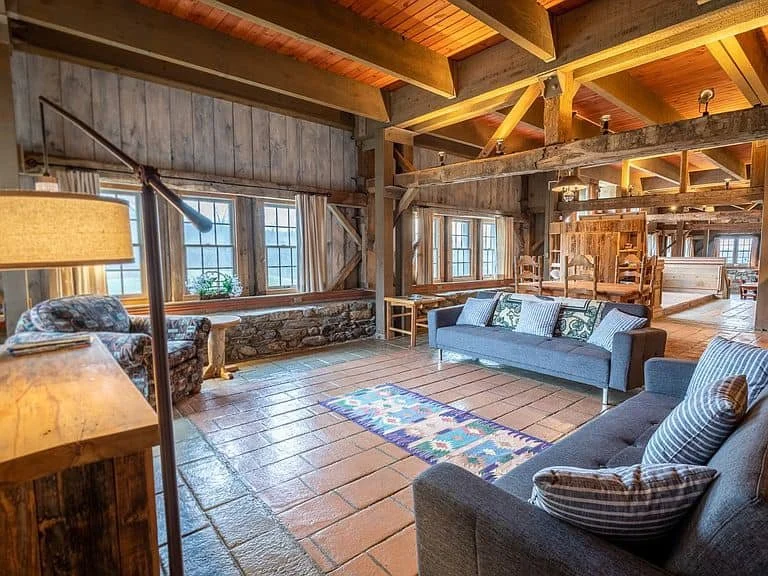Living room with rustic wooden interior, large windows, wooden beams, tiled floor, sofas, a small colorful rug, and a floor lamp.