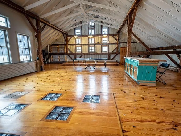 The rustic hayloft with natural wood floors, several windows, and exposed beams. Contains a ping pong table, a turquoise kitchen island, and a single chair.