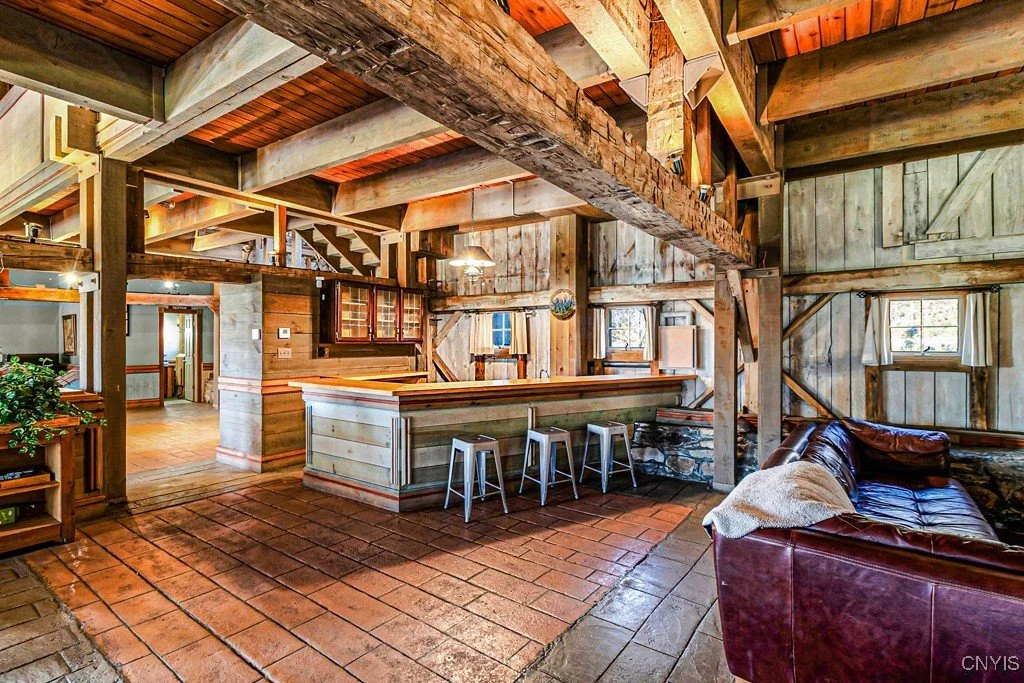 Interior of a rustic cabin living area with wooden beams, a bar counter with stools, brick flooring, a leather sofa, and small windows with curtains.