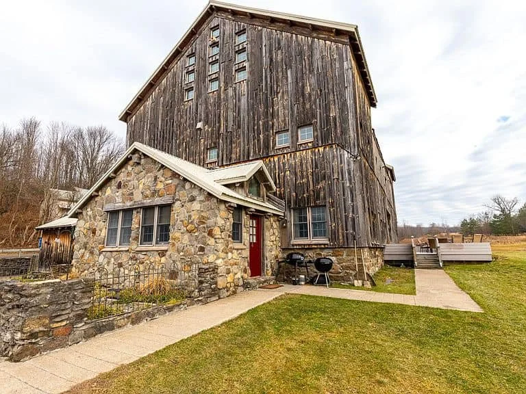 A rustic house with a stone foundation and wooden upper structure, featuring a red front door and small windows, surrounded by a grassy yard with a pathway and a backyard deck.
