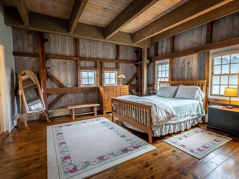 Bedroom with wooden walls, ceiling, and floors, featuring a bed with white bedding, a black nightstand with a lamp, a dresser with a lamp, a small rug, a mirror, and windows allowing natural light.