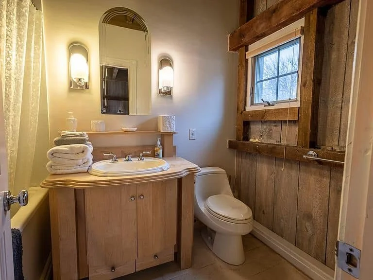Bathroom with wooden accents, a sink with a wooden cabinet, a mirror with two wall lights, a toilet, and a small window with a view of trees outside.