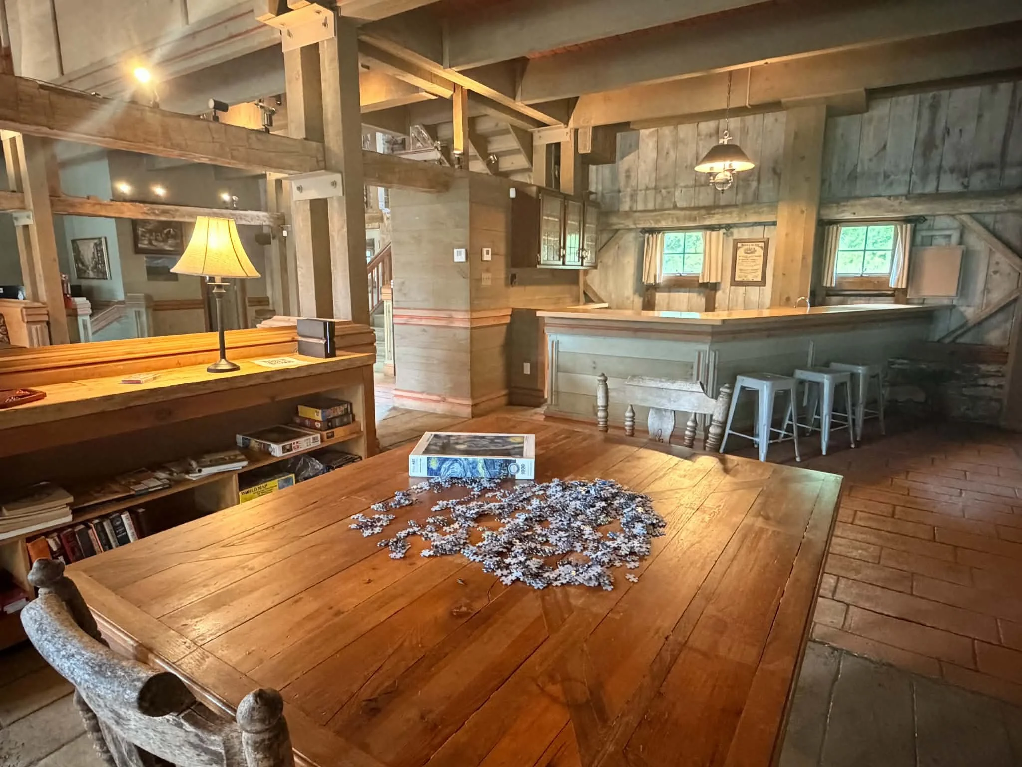 Interior of a rustic wooden house with a large wooden dining table in the foreground, a puzzle spread on it, and books underneath. To the left is an old wooden chair with a rolled armrest, and a lamp with a yellow shade. In the background, there are wooden beams, windows with curtains, and various furnishings, creating a cozy, cabin-like atmosphere.