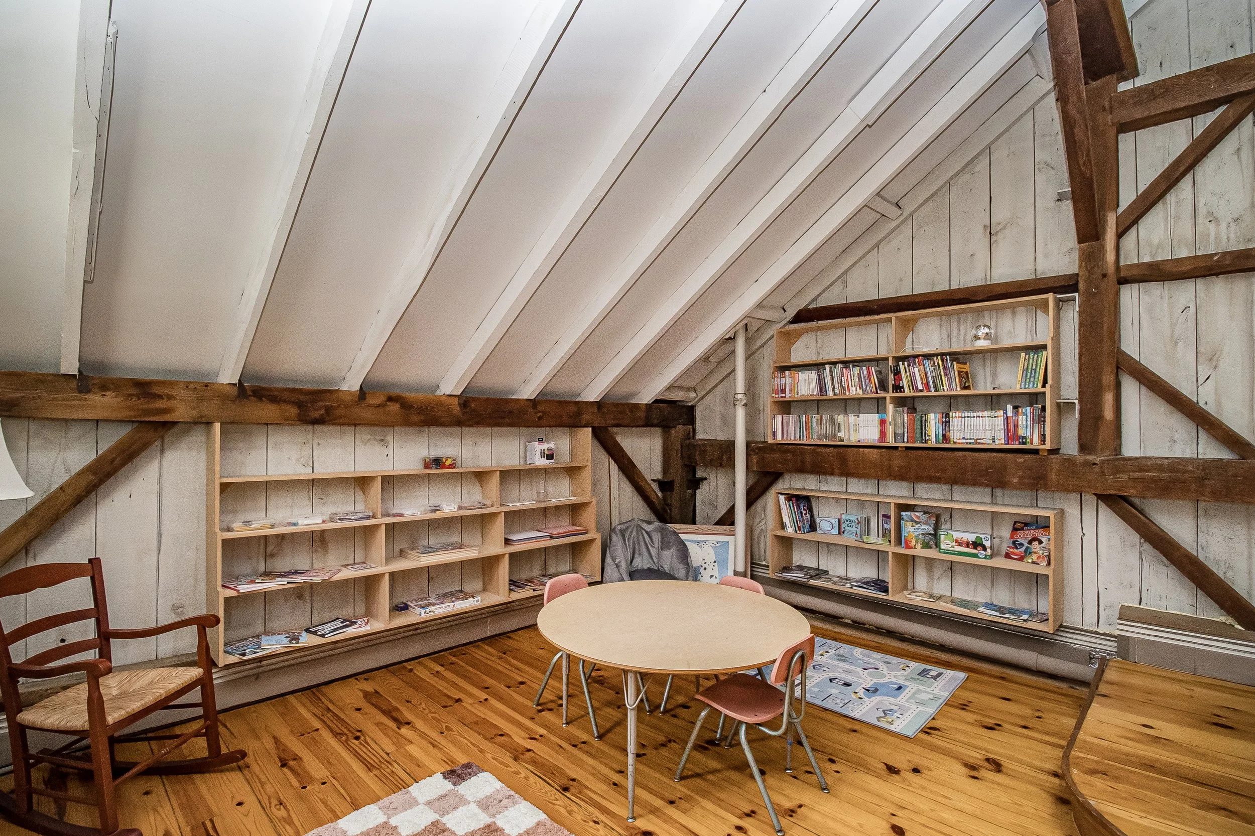 A cozy attic room with white painted wooden walls and ceiling, exposed wooden beams, and hardwood floors. There are bookshelves filled with books, a round table with chairs, and a patterned rug on the floor.