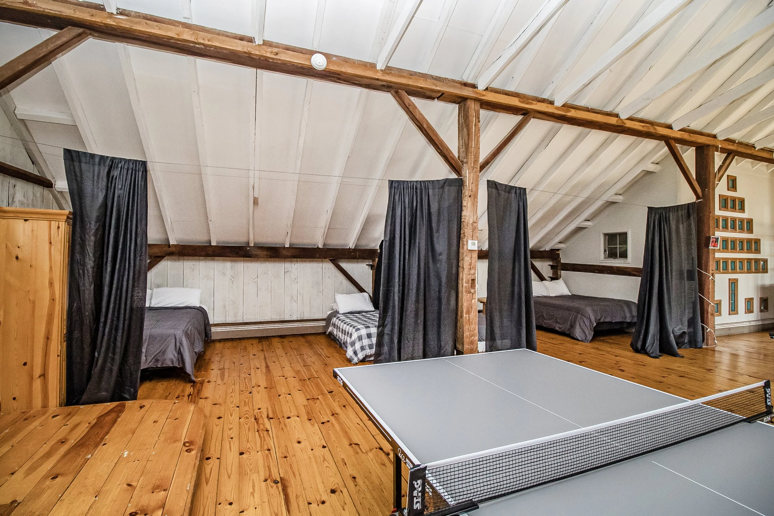 A spacious room in an attic with wooden floors, two beds with dark bedding, black curtains, and a ping pong table. The ceiling has white wooden panels with exposed beams and a smoke detector.