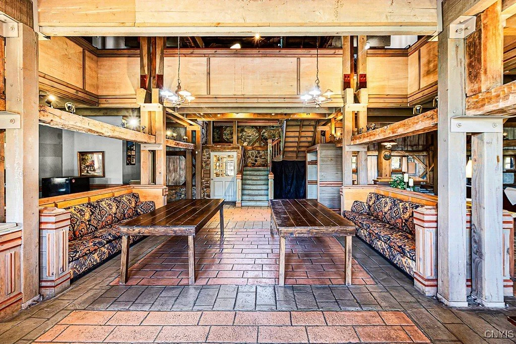 Interior of a rustic lodge with wooden beams, brick flooring, two long tables, and upholstered banquette seating.