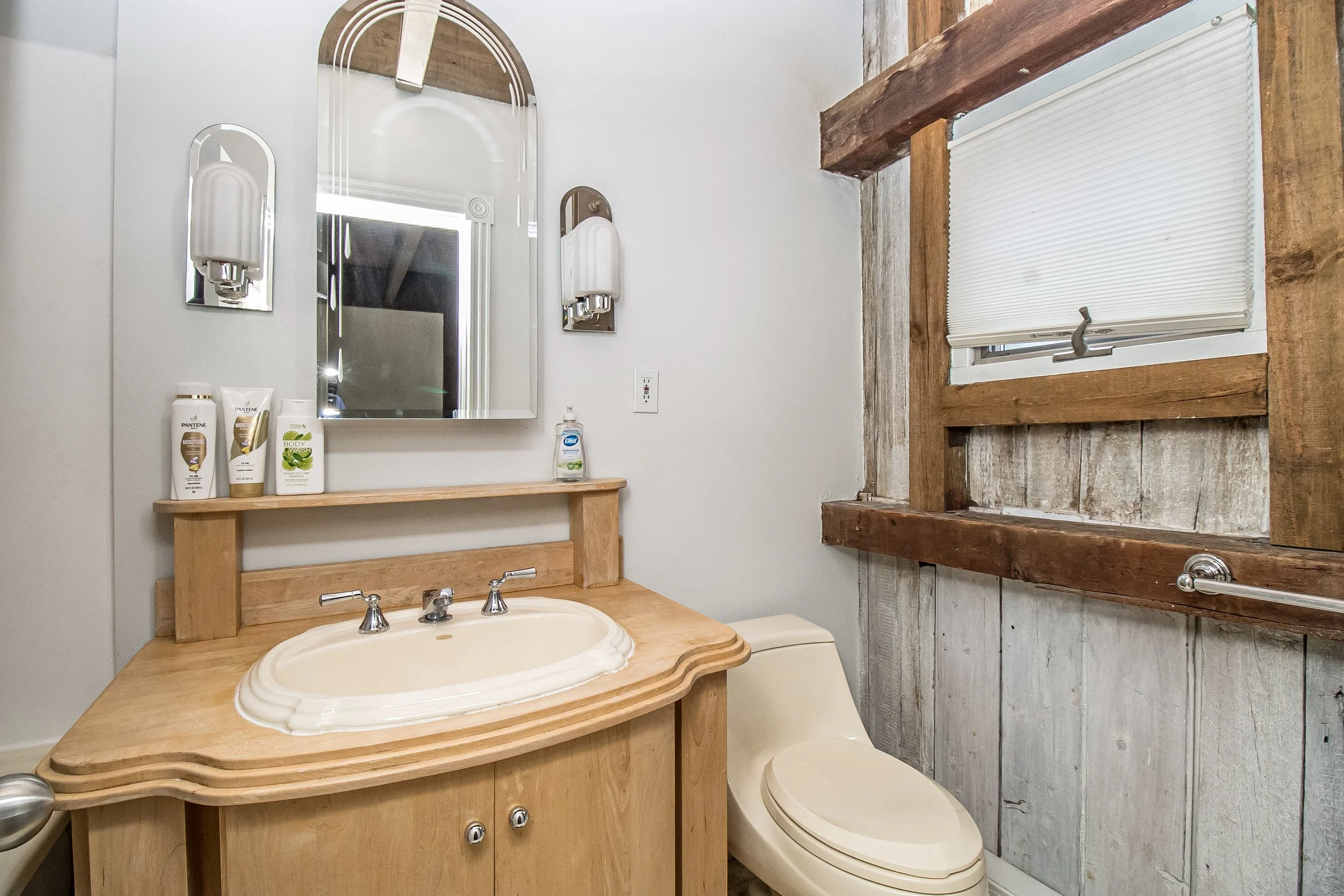 Small bathroom with a wooden vanity, a cream-colored sink, a mirror, and a decorated wall. There are toiletries on the shelf above the sink, including shampoo, body wash, and moisturizer. A window with a shutter lets in natural light. The wall and window frame are rustic, weathered wood.