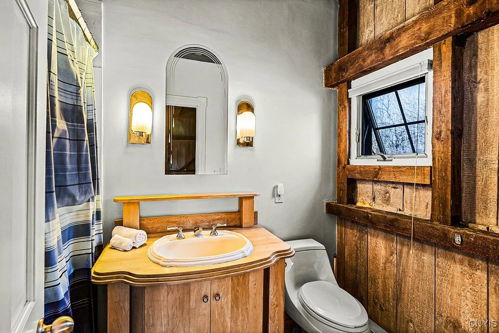 Rustic bathroom with a wooden vanity, a white toilet, a window with a wooden frame, and wood-paneled walls.