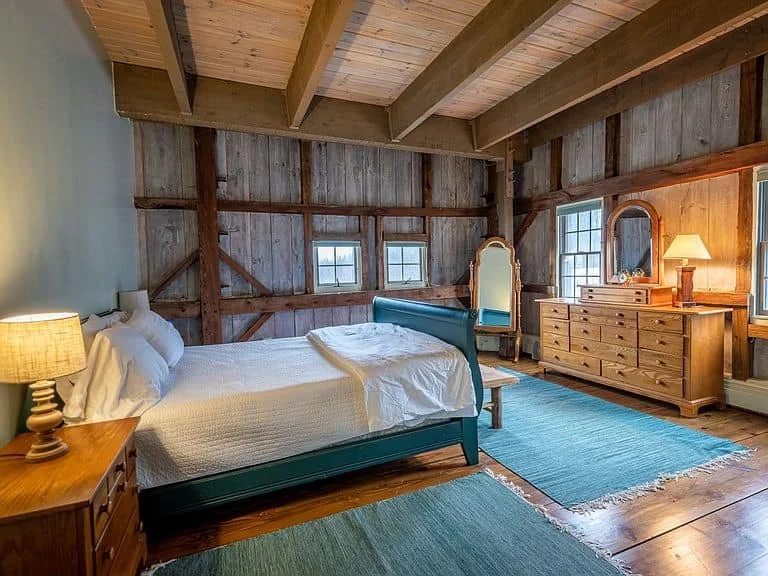 A rustic bedroom with a wooden beamed ceiling, a teal bed with white bedding, a wooden side table with a lamp, a dresser with a mirror, a vanity with a mirror, and windows letting in natural light.