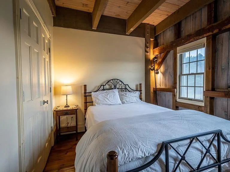 Cozy bedroom with wooden ceiling beams, a white bed with a metal headboard, a small wooden nightstand with a lamp, and a window showing trees outside.